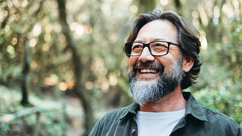 Man with salt and pepper beard and glasses smiling with defocused woods in background.